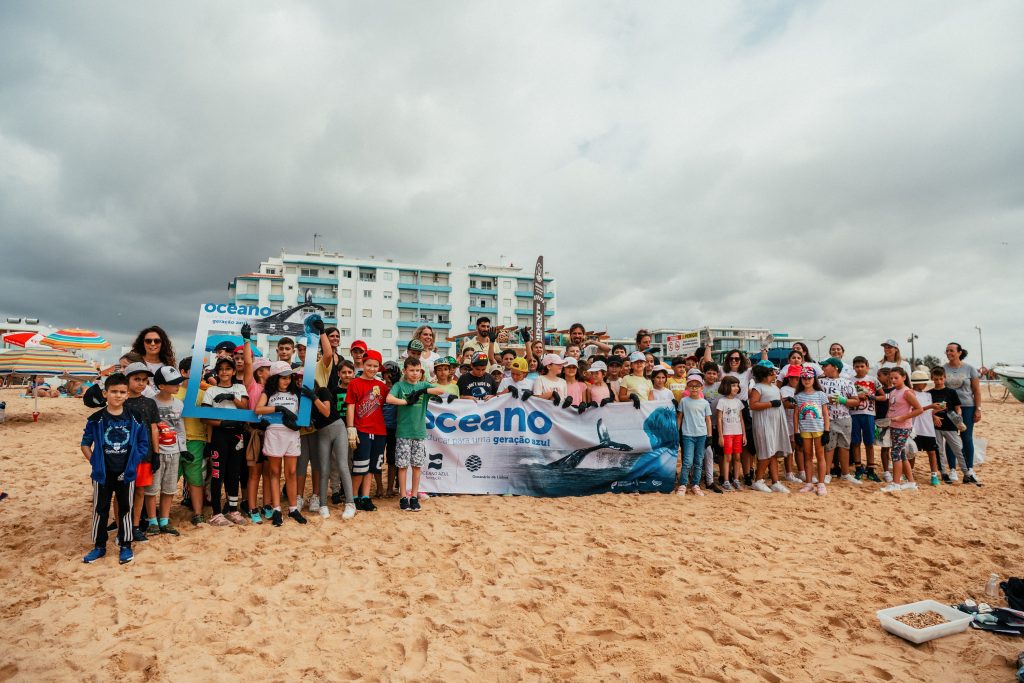 Group of children and adults smiling on a sandy beach, holding an "Oceano" banner. Cloudy sky and buildings in the background.