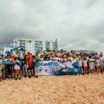 Group of children and adults smiling on a sandy beach, holding an "Oceano" banner. Cloudy sky and buildings in the background.