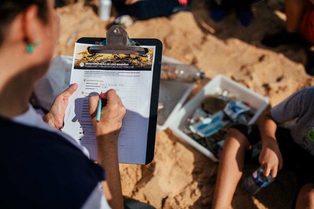 Person holding clipboard with form on a sandy beach, writing with a pen. Nearby, a box filled with collected trash. Scene of environmental cleanup.