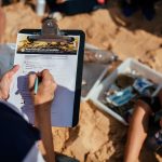 Person holding clipboard with form on a sandy beach, writing with a pen. Nearby, a box filled with collected trash. Scene of environmental cleanup.