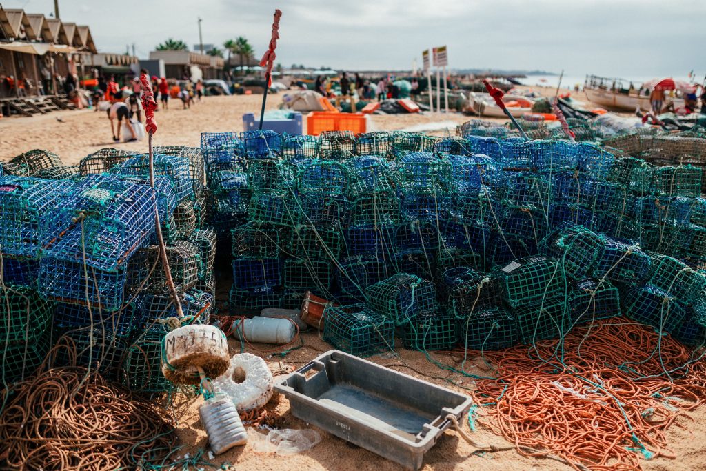 Stacks of blue lobster traps and orange ropes on a sandy beach. In the background, people walk near wooden huts and scattered boats under a cloudy sky.