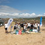 A group of people stand on a sandy beach, smiling after a cleanup effort. They are surrounded by collected trash bags and banners. The sky is clear and sunny.