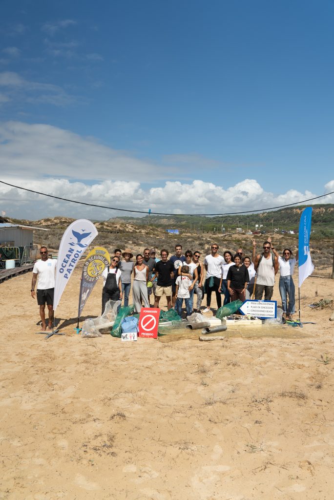 A group of people stand on a sandy beach, smiling after a cleanup effort. They are surrounded by collected trash bags and banners. The sky is clear and sunny.