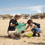 Two people clean a sandy beach, holding a green mesh bag of collected trash on a sunny beach. Clear blue sky and coastal landscape in the background.