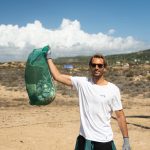 Man with sunglasses smiling, holding a green mesh bag of collected trash on a sunny beach. Clear blue sky and coastal landscape in the background.
