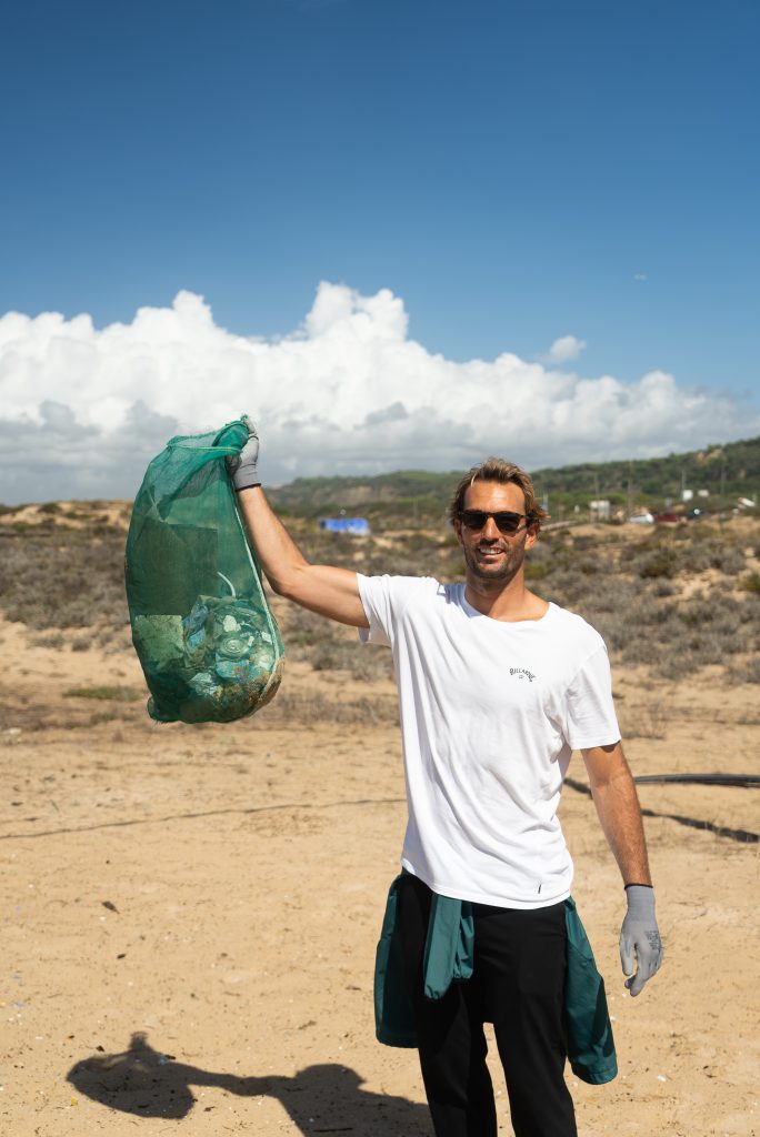 Man with sunglasses smiling, holding a green mesh bag of collected trash on a sunny beach. Clear blue sky and coastal landscape in the background.